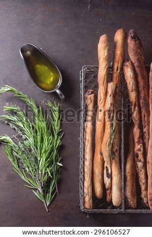 Fresh baked homemade grissini bread sticks in vintage metal grid box with olive oil and herbs rosemary and thym over dark surface. Top view.
