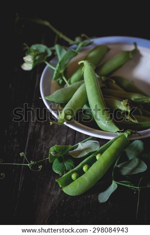 Heap of young green peas whole and broken with leaves and flower in vintage dinner plate over old wooden table. Dark rustic style. Natural day light.