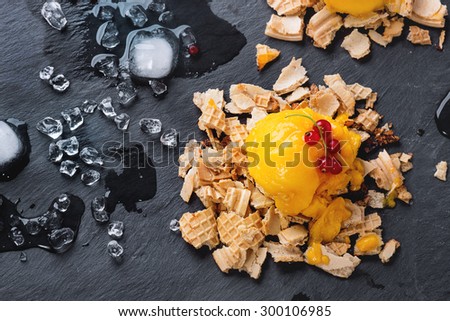 Mango sorbet ice cream, served on wafer crumbs with red currant berries and ice cubes over black slate. Top view.