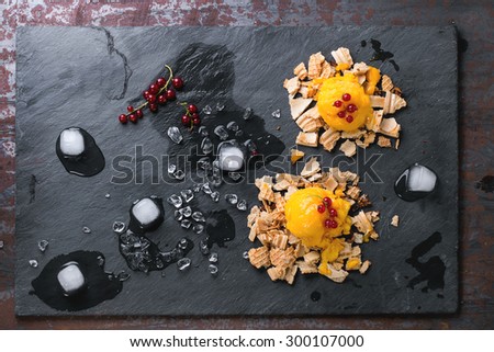 Mango sorbet ice cream, served on wafer crumbs with red currant berries and ice cubes over black slate. Old metal background. Top view.