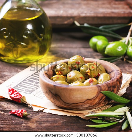 Green olives in olive wood bowl and bottle of olive oil served with chili peppers on old wooden table. Square image with selective focus
