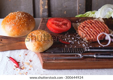 Ingredients for making homemade burger on wooden cutting board, served with meat fork and knife over White tablecloth. Dark rustic style.