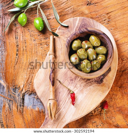 Green olives in olive wood bowl with chili pepper and olive\'s branch served on cutting board over wooden table. Top view. Square image