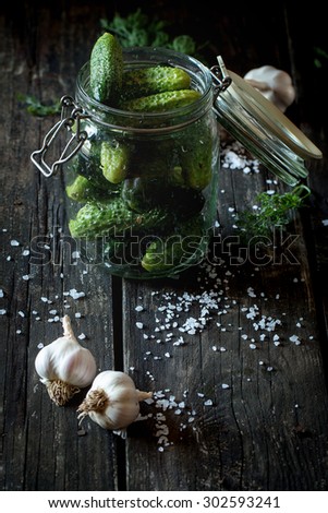 Glass jar with fresh low-salt pickled cucumbers. Over black wooden table with sprinkled sea salt and garlic. Dark rustic style. Natural day light