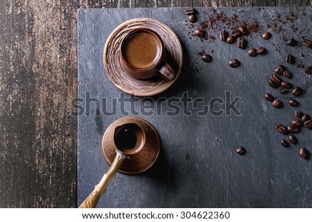 Brown ceramic cup of coffee, old copper cezve and coffee beans. Over black slate as background. With copy space at centre. Top view.