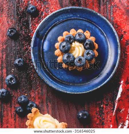 Top view on blueberry mini tarts served on blue ceramic plate over black and red wooden background. Square image