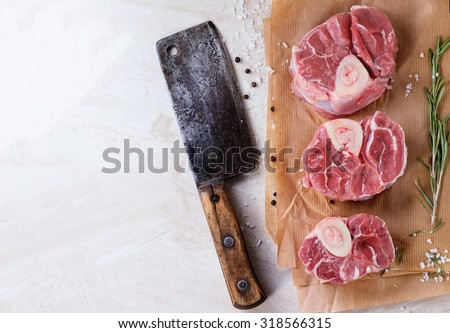 Raw osso buco meat on crumpled paper with salt, pepper and rosemary. With vintage backsword over white marble as background. Top view.