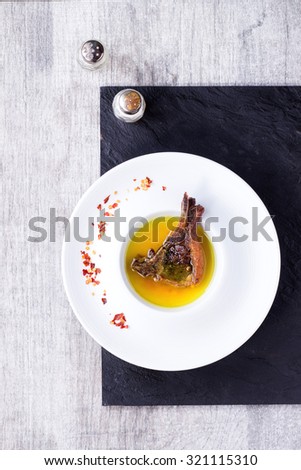 White ceramic plate with well done grilled lamb chop in yellow broth with chili pepper\'s flackes over black slate board over white wooden table. With vintage salt and pepper shakers near. Top view.
