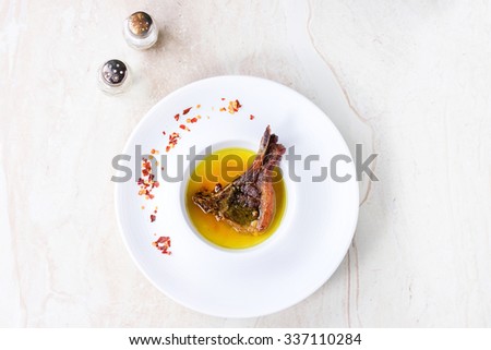White ceramic plate with well done grilled lamb chop in yellow broth with chili pepper\'s flackes over white marble table. Top view.