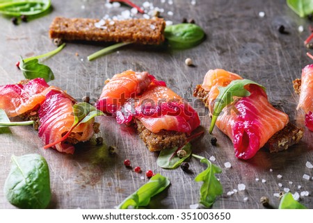 Sliced salmon filet, salted with beetroot juice, served on whole wheat toasts with salad leaves, sea salt and pepper over metal surface. Top view