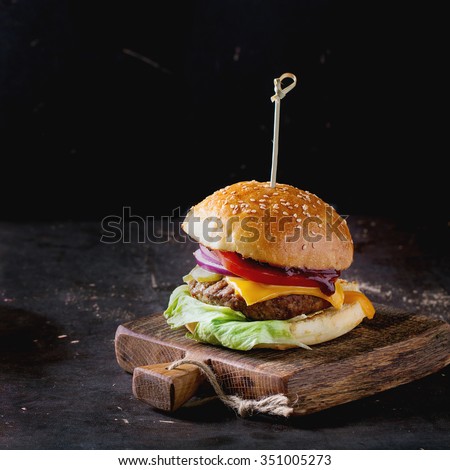 Fresh homemade burger on little wooden cutting board over dark background. Square image with selective focus