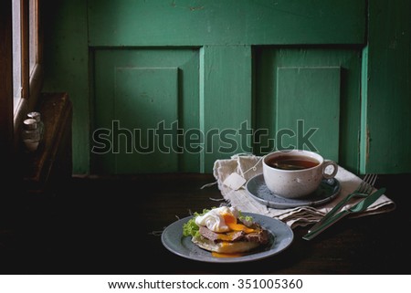 Cafe Breakfast with homemade sandwich with baked meat and soft-boiled egg and cup of hot pocket tea over dark wooden table near the window with turquoise wall at background. Dark rustic style.