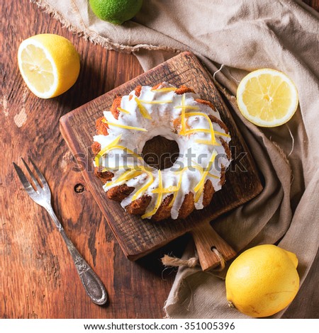Homemade cakes with white frosting and lemon zest, served with lemons and lime on small wooden cutting boards over wooden background with gray textile. Top view. Square image
