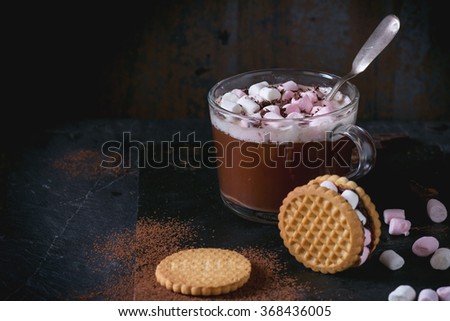 Glass cup of Hot chocolate with marshmallows, cookies stuffed by marshmallows, chopping chocolate and cocoa powder from sieve over double black slate surface. Dark rustic style.