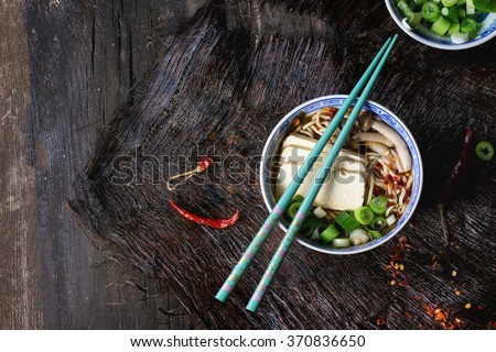 Chinese porcelain bowl of asian ramen soup with feta cheese, noodles, spring onion and mushrooms, served with turquoise chopsticks over old wooden table. Dark rustic style. Top view