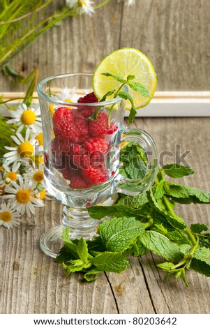 Still-life with fresh ripe raspberries in glass, bunch of camomile flowers and mint\'s leafes near mirror with reflection on old wooden table