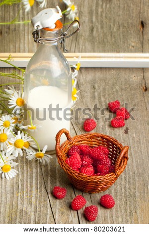 Still-life with basket of fresh ripe raspberries, bottle of milk and bunch of camomile flowers near mirror with reflex on old wooden table