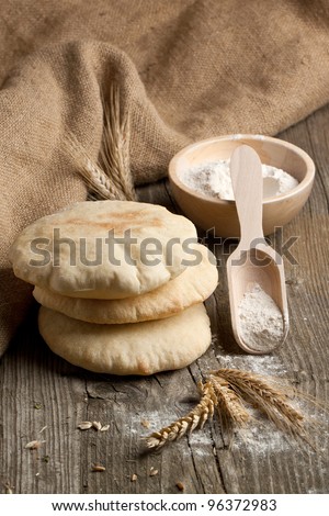 Three fresh pitas bread, ears of rye and bowl of flour on old wooden table