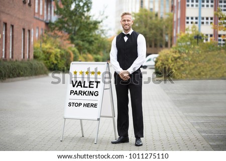 Portrait Of A Smiling Young Male Valet Standing Near Valet Parking Sign