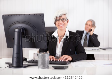 Smiling secretary or personal assistant sitting working at her desk with her boss visible on the telephone in the background