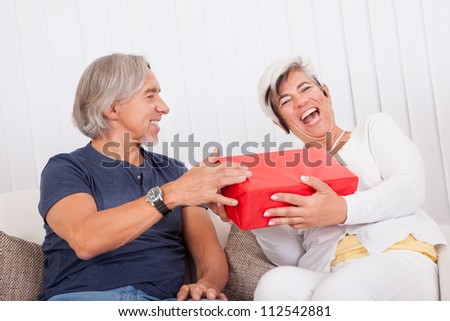 Laughing senior couple relaxing on a couch with the husband holding a red gift box