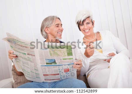 Senior couple sitting close together on a sofa sharing a newspaper as they catch up on the days news