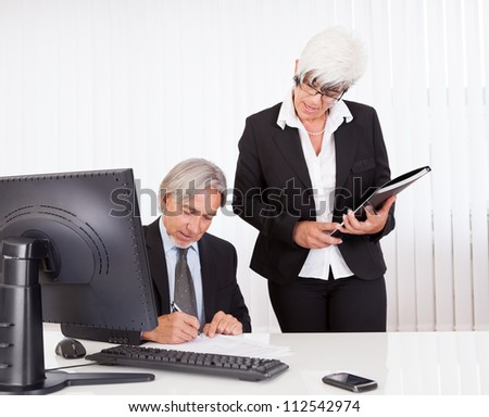 A senior business executive seated at his desk signs a document that has been brought to him by his secretary or personal assistant
