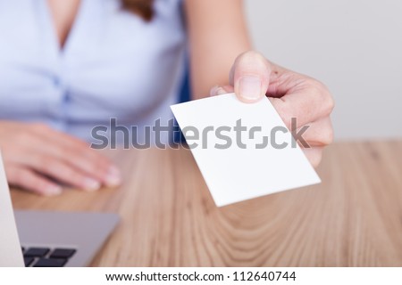 Young businesswoman sitting behind her laptop holds out her hand offering her card with focus to her face