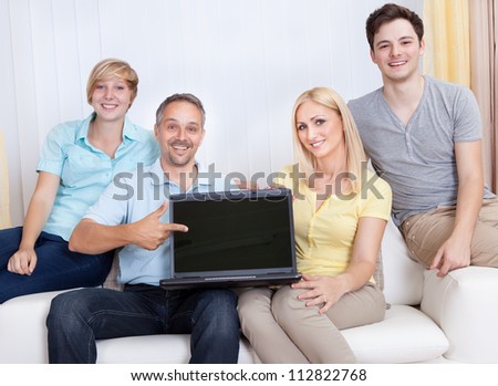 Smiling father surrounded by his happy teenage family pointing to a blank laptop screen for your advertising and marketing