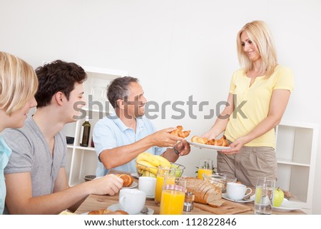 Young healthy family seated at the table being served croissants for breakfast by the smiling mother
