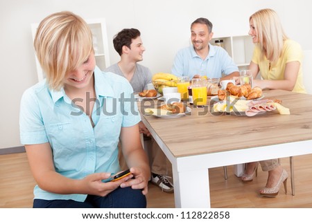 Teenager texting on her mobile phone in the kitchen while the rest of the family enjoy breakfast