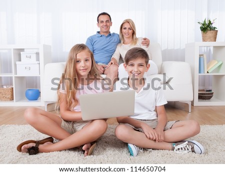 Children On The Carpet Using Tablet And Laptop With Parents Behind Them At Home
