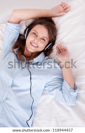 Woman relaxing on her bed in a casual blue shirt wearing stereo headphones bed listening to music