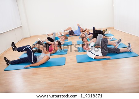 Large group of diverse people exercising in a gym class lying on blue mats doing leg raising and twisting exercises