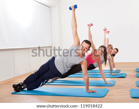 Large group of people working out in a gym balanced on one hand on their mats while raising their other arm with a dumbbell