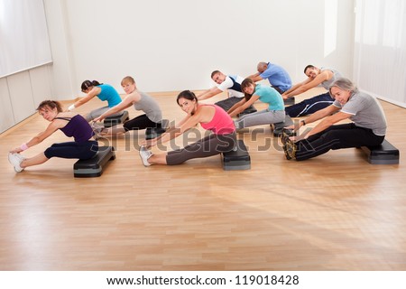 Large group of people doing stretching exercises in a gym sitting on boards reaching for their feet which are extended in front of them