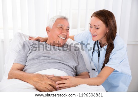 Nurse Looking At Happy Male Patient Lying On Bed In Clinic