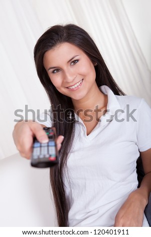 Smiling woman sitting on a couch with a remote control in her hands with copyspace over white curtains