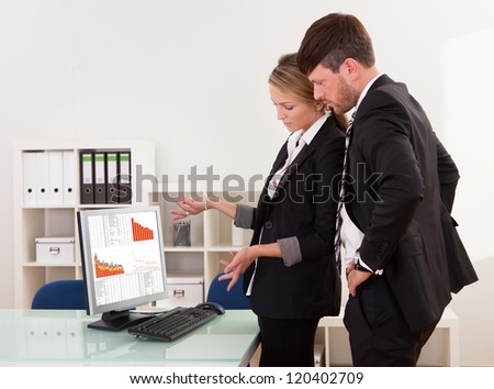 Two professional young business colleagues stand in an office looking at a computer with dropping descending graphs