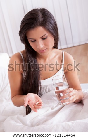 Ill woman in bed taking medication sitting up holding a glass of water looking at a tablet in her hand