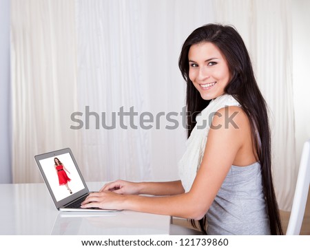 Happy woman sitting at her desk shopping online browsing colored clothing catalogues of models in dresses