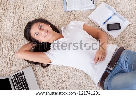 Overhead view of a young woman lying on her back on a carpet alongside her laptop daydreaming