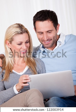 Beautiful young couple with lovely smiles sitting side by side on a sofa looking at a tablet together