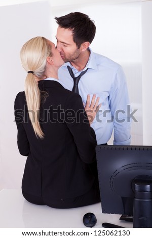 A businessman embraces a female colleague who is sitting on a desktop as mutual passion leads to an affair or love in the workplace