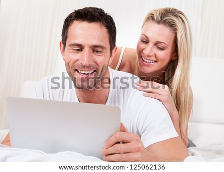 Young couple reading a laptop screen with the husband lying on his stomach on a bed and his wife leaning on him looking over his shoulder