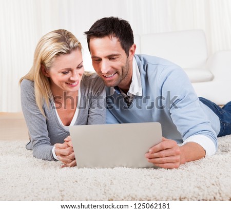 Attractive casual couple lying on their stomachs on a carpet with a laptop reading the information on the screen together
