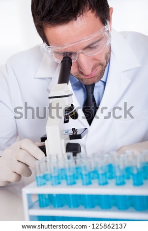 Male pathologist or lab technician using a microscope with a rack of test tubes containing blue liquid in front of him