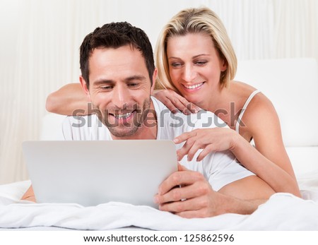 Young couple reading a laptop screen with the husband lying on his stomach on a bed and his wife leaning on him looking over his shoulder