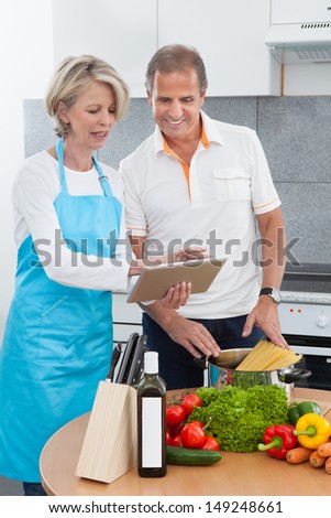 Mature Man And Woman Looking At Recipe Tablet While Cooking In Kitchen