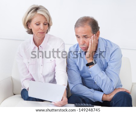 Portrait Of Shocked Couple Sitting On Couch Looking At Bill